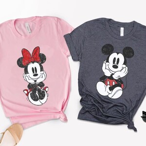 Pode incluir: Duas camisetas com personagens da Disney. Uma &eacute; rosa claro com Minnie Mouse usando um la&ccedil;o vermelho. A outra &eacute; cinza escuro com Mickey Mouse. Ambas as camisas t&ecirc;m um design gr&aacute;fico desgastado.