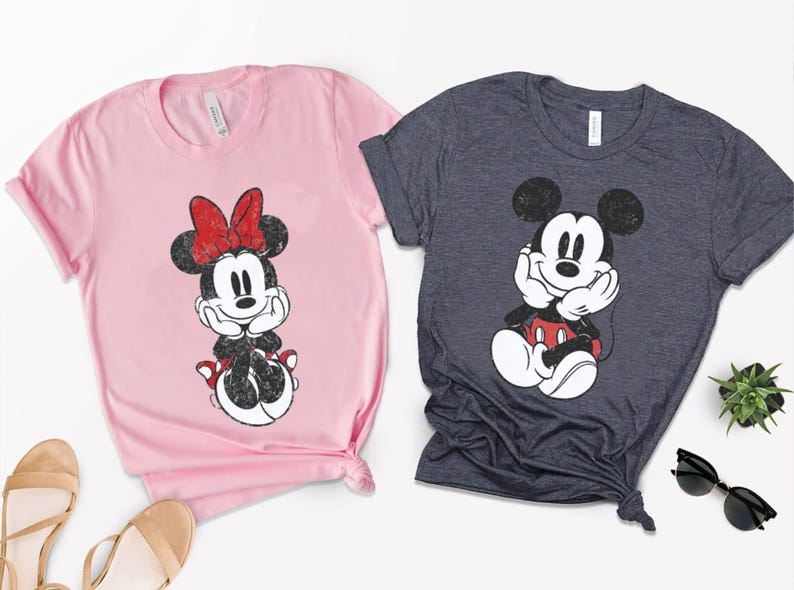 Pode incluir: Duas camisetas com personagens da Disney. Uma &eacute; rosa claro com Minnie Mouse usando um la&ccedil;o vermelho. A outra &eacute; cinza escuro com Mickey Mouse. Ambas as camisas t&ecirc;m um design gr&aacute;fico desgastado.