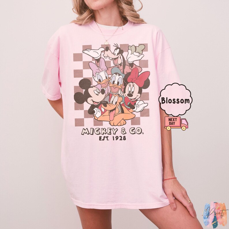 May include: A light pink t-shirt featuring a graphic of Mickey Mouse and friends. The design includes Mickey, Minnie, Donald, Daisy, Goofy, and Pluto, set against a brown and white checkered background. The text "Mickey & Co. Est. 1928" is printed below.