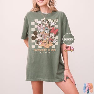 May include: Green t-shirt featuring a graphic of Mickey Mouse and friends. The design includes Mickey, Minnie, Donald, Daisy, Goofy, and Pluto, with the text "Mickey & Co. Est. 1928" in a vintage style. The shirt has a relaxed fit.