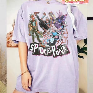 May include: A light purple t-shirt featuring a graphic of several Spider-Man characters in punk rock attire. The words "SPIDER-PUNK" are printed in a black, stylized font across the bottom of the graphic. The shirt has short sleeves and a crew neck.