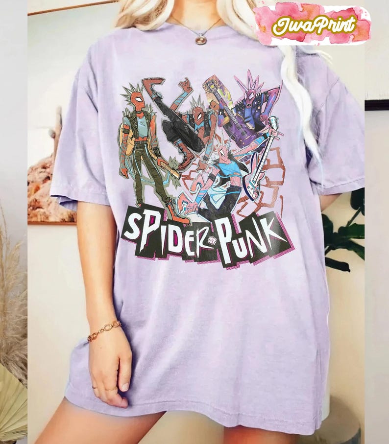 May include: A light purple t-shirt featuring a graphic of several Spider-Man characters in punk rock attire. The words "SPIDER-PUNK" are printed in a black, stylized font across the bottom of the graphic. The shirt has short sleeves and a crew neck.