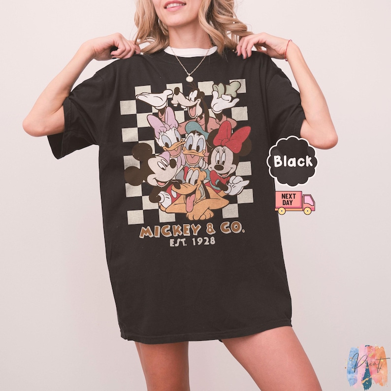 May include: Black t-shirt featuring a group of Disney characters on a checkered background. The shirt has the text "Mickey & Co. Est. 1928" printed on it. The shirt is a classic fit.