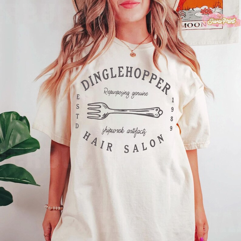 Puede incluir: Camiseta color crema con un gr&aacute;fico negro que dice "DINGLEHOPPER HAIR SALON" con una ilustraci&oacute;n de tenedor. El texto incluye "Repurposing genuine" y "shipwrek artifacts". La camiseta tambi&eacute;n tiene las letras "ESTD" y los n&uacute;meros "1989".