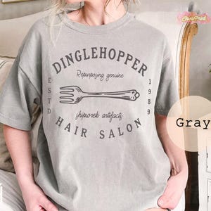 Puede incluir: Camiseta gris con el texto "DINGLEHOPPER HAIR SALON" en un dise&ntilde;o circular, con una ilustraci&oacute;n de tenedor. La camiseta tambi&eacute;n incluye el texto "Repurposing genuine" y "shipwrek artifacts".