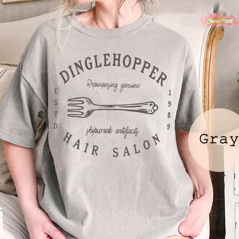 Puede incluir: Camiseta gris con el texto "DINGLEHOPPER HAIR SALON" en un dise&ntilde;o circular, con una ilustraci&oacute;n de tenedor. La camiseta tambi&eacute;n incluye el texto "Repurposing genuine" y "shipwrek artifacts".