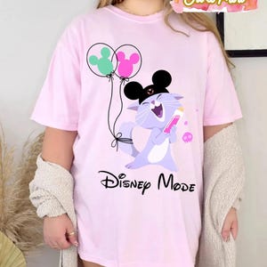Puede incluir: Camiseta rosa claro con un gato de dibujos animados con orejas de Mickey y un objeto rosa. El gato se ríe. Dos globos con forma de orejas de Mickey flotan arriba. El texto "Disney Mode" está impreso debajo.