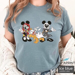Puede incluir: Camiseta azul hielo de color confort con un dibujo animado de Minnie Mouse, Pluto y Jack Skellington con disfraces de Halloween. Los personajes son de The Nightmare Before Christmas. La camiseta tiene el texto "JW APRINT" y "ice blue comfort color".
