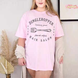 Puede incluir: Camiseta rosa claro con el texto "DINGLEHOPPER HAIR SALON" en un dise&ntilde;o circular. El dise&ntilde;o incluye una ilustraci&oacute;n de tenedor y las palabras "Repurposing genuine" y "shipwrek artifacts". La camiseta tiene mangas cortas.