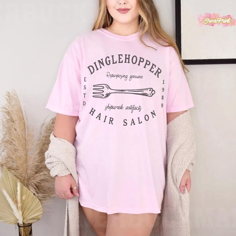 Puede incluir: Camiseta rosa claro con el texto "DINGLEHOPPER HAIR SALON" en un dise&ntilde;o circular. El dise&ntilde;o incluye una ilustraci&oacute;n de tenedor y las palabras "Repurposing genuine" y "shipwrek artifacts". La camiseta tiene mangas cortas.