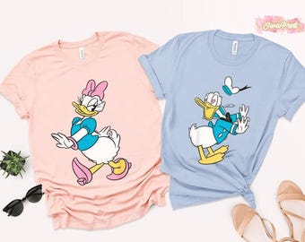 May include: Two t-shirts, one pale pink and one pale blue, each with a cartoon character. The pink shirt features Daisy Duck, and the blue shirt features Donald Duck. Both characters are depicted in motion, with bright colours and bold outlines.