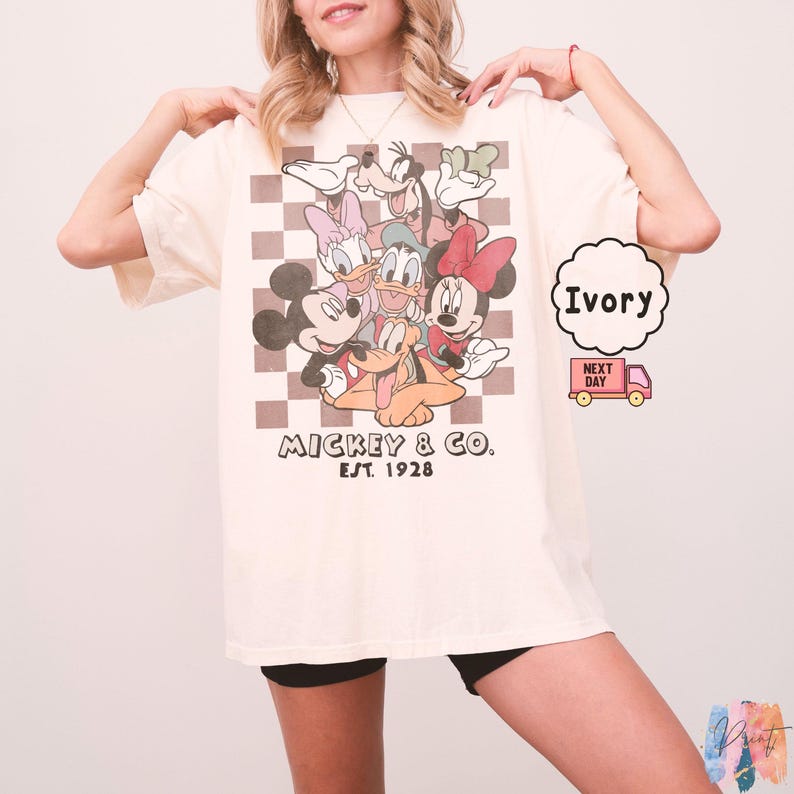 May include: Ivory-colored t-shirt featuring a graphic of Mickey Mouse, Minnie Mouse, Donald Duck, Daisy Duck, Goofy, and Pluto. The graphic is set against a brown and white checkered background. The text "Mickey & Co. Est. 1928" is printed below the characters.