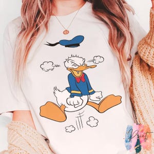 May include: White t-shirt featuring a cartoon image of Donald Duck in a blue sailor suit, red bow tie, and blue hat. The character is depicted with an angry expression, jumping with his feet in the air. The design includes cartoon clouds.