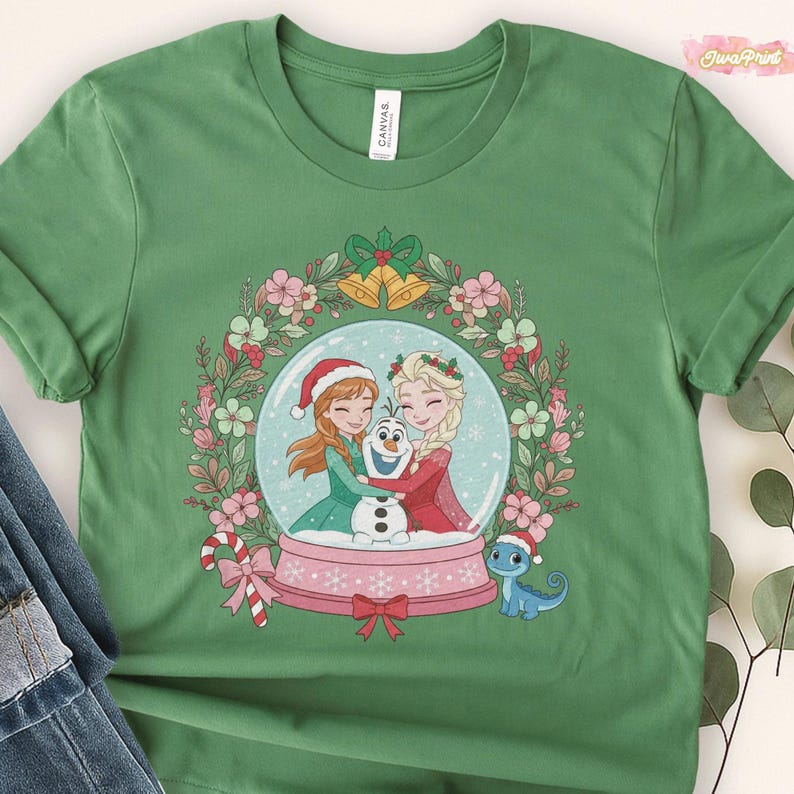 May include: Green t-shirt featuring a snow globe design with two cartoon characters hugging Olaf the snowman. The snow globe is surrounded by a floral wreath, bells, and a candy cane. A small blue dinosaur wearing a Santa hat is also present.