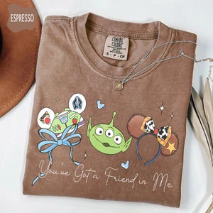 May include: Espresso-colored t-shirt featuring a graphic of Toy Story characters, including Buzz Lightyear, an alien, and mouse ears. The shirt has the phrase "You've Got a Friend in Me" and is paired with a silver necklace.