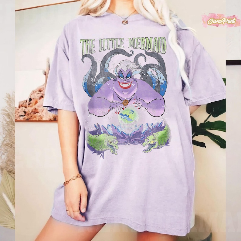 May include: A light purple t-shirt featuring a graphic of Ursula from The Little Mermaid. The design includes the text "THE LITTLE MERMAID" in green, Ursula's face, tentacles, and two green eels. The shirt is a relaxed fit.