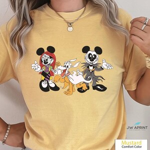 Puede incluir: Camiseta de color mostaza con un gr&aacute;fico de dibujos animados de Minnie Mouse, Pluto y Jack Skellington. Minnie lleva una peluca roja y un atuendo morado y negro. Pluto tiene una s&aacute;bana blanca. Jack lleva un traje negro y gris.