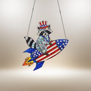 May include: A stained glass suncatcher featuring a raccoon wearing a patriotic Uncle Sam hat, riding a rocket ship. The rocket has red and white stripes and blue stars, with a flame of yellow, orange, and blue. The suncatcher hangs from a chain.
