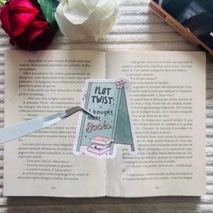 Op de afbeelding: Een sticker met de tekst "PLOT TWIST: I bought MORE Books" en een illustratie van een stapel boeken. De sticker staat op een open boek met een rode roos en een witte roos op de achtergrond.