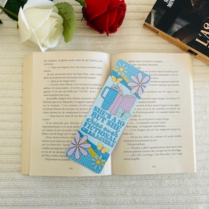 May include: A patterned bookmark with the text "SHE'S A 10 BUT SHE CHIES OVER FICTIONAL CHARACTERS" is inserted in a book. The bookmark has a blue background with floral and butterfly designs, and illustrations of a mug and books.