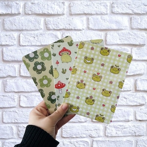 以下が含まれることがあります： 手には、3枚の装飾カードが持たれています。カードには、緑色の花、キノコ、笑顔のカエルが描かれています。背景は、白いレンガの壁です。