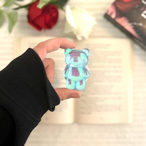 May include: A small, iridescent blue and purple teddy bear figurine. The bear has a geometric design and is held in front of an open book, with red and white roses in the background.