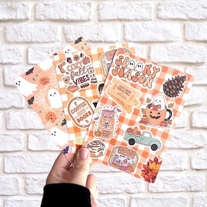 Peut inclure: Une main tient une pile d'autocollants sur le thème de l'automne, devant un mur de briques blanches. Les autocollants présentent des fantômes, des citrouilles et des phrases comme "Spooky Season" et "Cozy Fall Vibes" en orange et blanc.