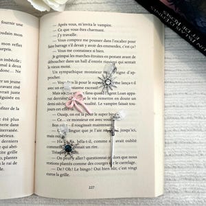 Op de afbeelding: Decoratieve bladwijzers met verschillende bedels, waaronder een zilveren sneeuwvlok, een roze strik en een donkerblauwe edelsteen. De bladwijzers liggen op de pagina's van een open boek, met een witte roos op de achtergrond.