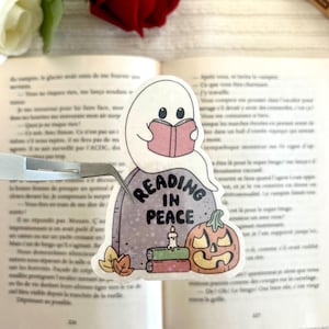 Peut inclure: Un autocollant fantaisiste représentant un fantôme lisant un livre, posé sur une pierre tombale avec l'inscription "READING IN PEACE". Une citrouille d'Halloween et des livres sont également présents. L'autocollant est posé sur un livre ouvert.