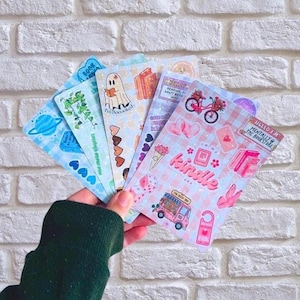 May include: A hand holding a selection of colourful stickers with diverse designs. The stickers depict books, planets, hearts, and text such as "Kindle" and "Hello I'm Mentally @ The Bookstore". The backdrop is a white brick wall.