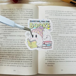 Peut inclure: Autocollant représentant un fantôme mignon avec un bonnet rose et blanc, lisant un livre. Le texte "Ghosting You For Books" et une pile de livres sont également présents.