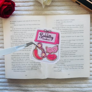 Peut inclure: Un autocollant transparent avec une boîte rose portant l'inscription "Smatty Cookies" et des illustrations d'un piment, d'une clé, d'un masque et de lèvres. L'autocollant est posé sur un livre ouvert.