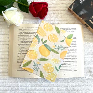 Pode incluir: Um marcador de livros com um padrão de limões, folhas e pequenas flores brancas. Está colocado em um livro aberto, com uma rosa vermelha e uma rosa branca ao fundo.