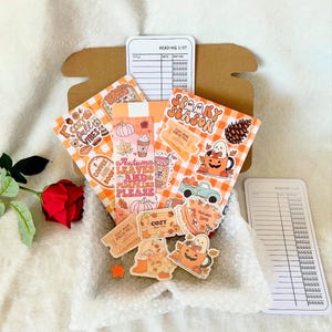 Op de afbeelding: Een open kartonnen doos gevuld met herfstthema stickers en kaarten. De stickers tonen pompoenen, koffiekopjes en de zinnen "Autumn Leaves and Pumpkins Please" en "Spooky Season". Een rode roos is zichtbaar.