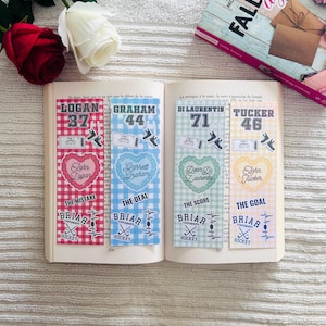 Op de afbeelding: Open boek met vier decoratieve bladwijzers. Elke bladwijzer heeft een naam, nummer en hockeyafbeeldingen. De bladwijzers zijn rood, blauw, groen en geel, met "Briar Hockey" onderaan. Een boek en bloemen staan op de achtergrond.