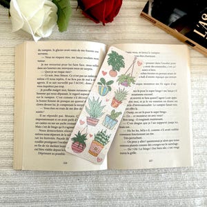 Puede incluir: Un marcapáginas decorativo con un patrón repetido de plantas en macetas y corazones de colores. El marcapáginas está colocado en un libro abierto con texto visible en las páginas.