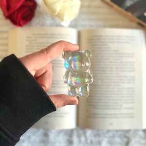 Può includere: Una statuetta a forma di orsetto trasparente e iridescente è tenuta davanti a un libro aperto. L'orsetto ha una finitura liscia e lucida e riflette un arcobaleno di colori. Una rosa rossa e una rosa bianca sono sullo sfondo.