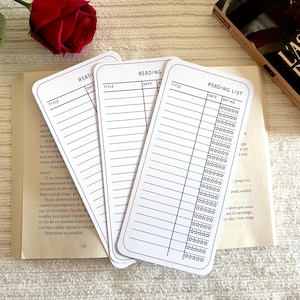 Könnte beinhalten: Drei weiße Lesezeichen mit schwarzen Texten, die Leselisten enthalten, mit Abschnitten für Titel, Datum und Bewertung. Die Lesezeichen liegen auf einem offenen Buch, mit einer roten Rose im Hintergrund.