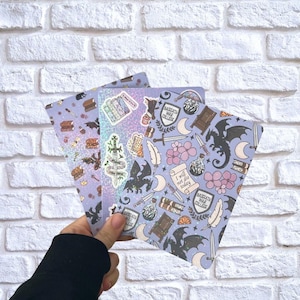 Puede incluir: Tres cuadernos de temática fantástica con varias ilustraciones. Los cuadernos presentan imágenes de dragones, libros, espadas y el texto "I will not die today" y "Basgiath War College" sobre un fondo lila claro.