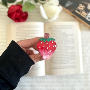 Peut inclure: Une barrette à cheveux en forme de fraise rouge et rose, avec des détails de graines blanches et une tige verte. La barrette est tenue devant un livre ouvert, avec une rose rouge et une rose blanche en arrière-plan.