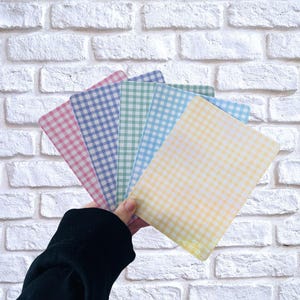 May include: A hand displays a fan of pastel-coloured, gingham-patterned paper sheets. The sheets are pink, blue, green, light blue, and yellow. The background is a white brick wall.