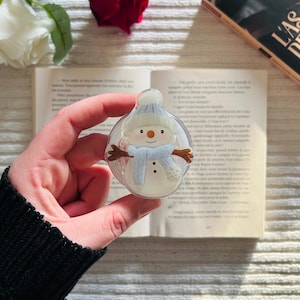 Puede incluir: Adorno navideño redondo y transparente con un diseño de muñeco de nieve. El muñeco de nieve tiene una bufanda y un gorro azul claro, brazos marrones y una nariz de zanahoria. El adorno se sostiene frente a un libro abierto y una rosa roja.