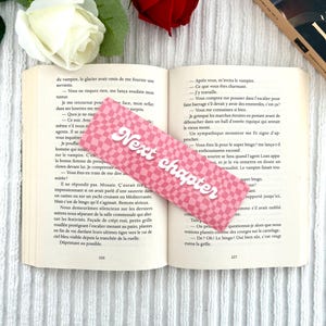 Peut inclure: Un marque-page rose et blanc à carreaux avec l'inscription "Next chapter" en blanc, posé sur un livre ouvert. Une rose rouge et une rose blanche sont visibles en arrière-plan.