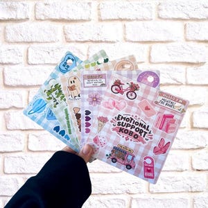May include: A hand holding a collection of decorative stickers. The stickers display images of a ghost, a bicycle with flowers, hearts, and text that says "Emotional Support Kobo" and "Hello I'm Reading". The backdrop is a white brick wall.
