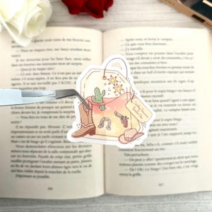 Può includere: Un adesivo a forma di bustina di tè a tema cowboy. L'adesivo presenta un cactus, stivali da cowboy, un ferro di cavallo, una pistola e un cappello da cowboy. L'etichetta recita "Cowboy Dreams, Southern Tea".