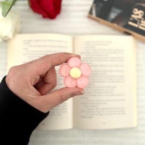 Op de afbeelding: Een roze bloemvormig accessoire met een gele kern wordt voor een open boek gehouden. De bloem heeft een zacht, afgerond ontwerp. Een rode roos en een boek zijn zichtbaar op de achtergrond.