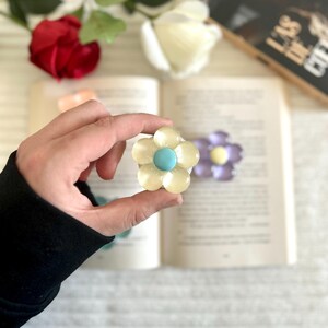 Peut inclure: Marque-page en forme de fleur avec des pétales jaune clair et un centre turquoise. Une main tient le marque-page devant un livre ouvert. Un deuxième marque-page en forme de fleur violette avec un centre jaune est également visible.