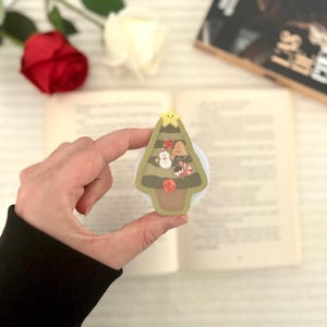 Peut inclure: Un support de téléphone en forme de sapin de Noël, orné d'une étoile, d'un bonhomme de neige, d'un bonhomme en pain d'épices et de cannes en sucre. L'arbre vert a des détails marron. Le support est tenu devant un fond flou de livre et de fleurs.