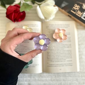 Könnte beinhalten: Nahaufnahme eines lila blütenförmigen Lesezeichens mit gelbem Zentrum, das in einer Hand gehalten wird. Ein weiteres pfirsichfarbenes Blütenlesezeichen ist auf einem offenen Buch zu sehen. Eine rote Rose und eine weiße Rose sind im Hintergrund.