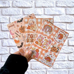 May include: Three decorative stickers with a fall theme are held in front of a white brick wall. The stickers feature pumpkins, bats, and the text "Spooky", "Boo to Yo", "In my cozy girl era", and "Pumpkin Season".
